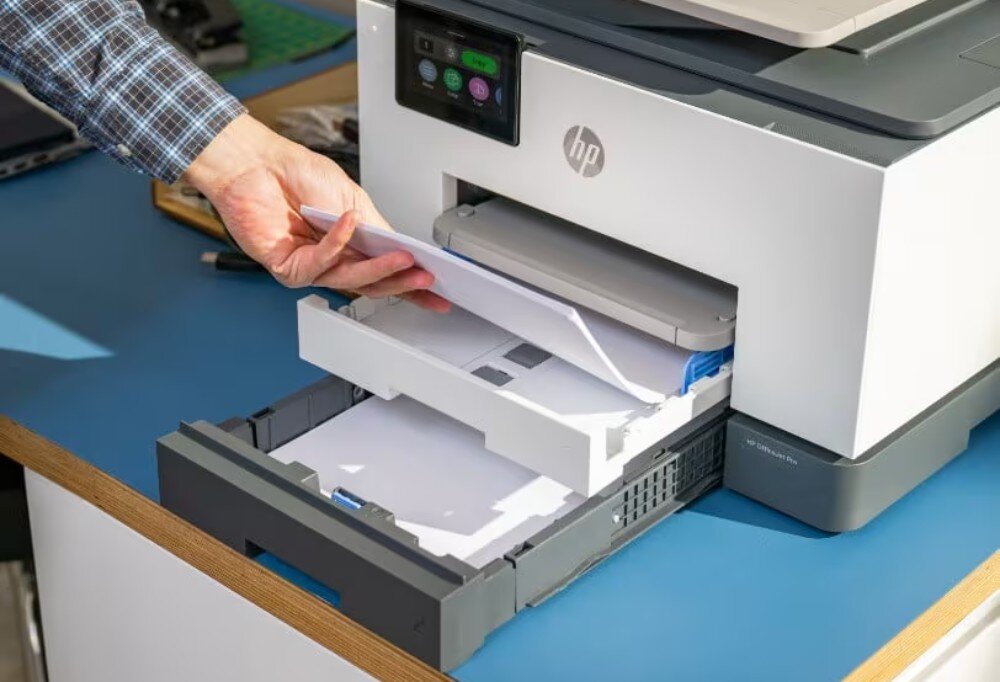 Osoba wkłada papier do szuflady drukarki wielofunkcyjnej z widocznym logo HP. Drukarka znajduje się na niebieskim blacie, a na panelu sterowania widoczne są przyciski i wyświetlacz.