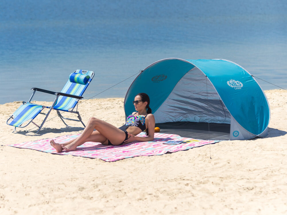 Na plaży znajduje się turkusowo-szary namiot z logo \'NILS CAMP\', obok którego leży osoba na ręczniku. W tle widać leżak w niebiesko-białe paski oraz wodę.