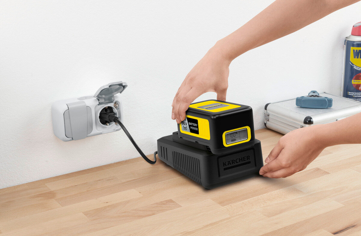 Osoba umieszcza żółto-czarną baterię na czarnej ładowarce z logo KARCHER, podłączonej do gniazdka elektrycznego. Na blacie obok znajduje się metalowa walizka i puszka z napisem \'WD-40\'.