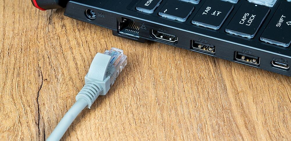 Szary kabel RJ-45 leży na drewnianej powierzchni obok klawiatury z widocznymi portami USB, HDMI i Ethernet.