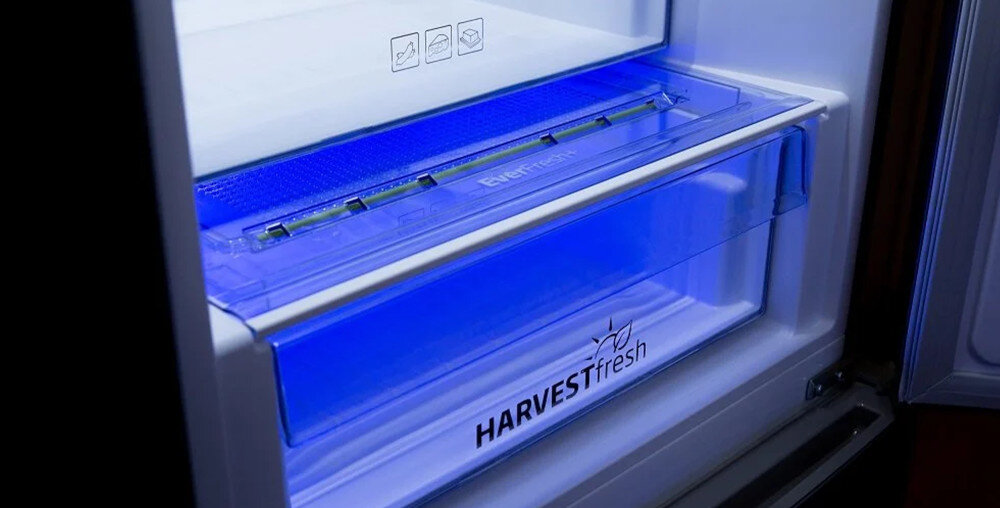 Wnętrze lodówki z niebieskim oświetleniem, widoczna szuflada z napisem \'HARVESTfresh\'.