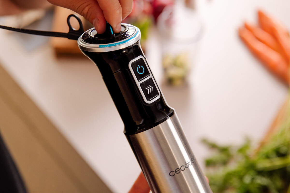 Osoba trzyma blender ręczny z widocznymi przyciskami i logo \'cecotec\'. W tle znajdują się rozmyte warzywa, w tym marchewki i zioła.