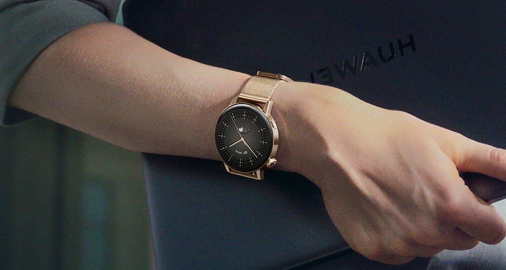 Osoba trzymająca ciemny przedmiot, na nadgarstku ma złoty smartwatch z okrągłą tarczą. Na przedmiocie widoczny jest napis \'HUAWEI\'.