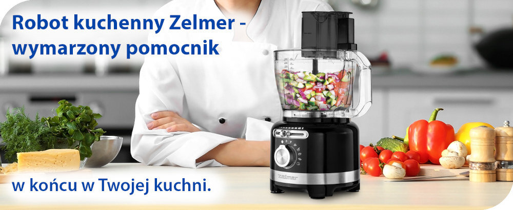 Na blacie kuchennym stoi czarny robot kuchenny z przezroczystym pojemnikiem wypełnionym pokrojonymi warzywami. Obok widoczne są świeże warzywa, zioła i ser, a w tle osoba w białym stroju kucharskim.