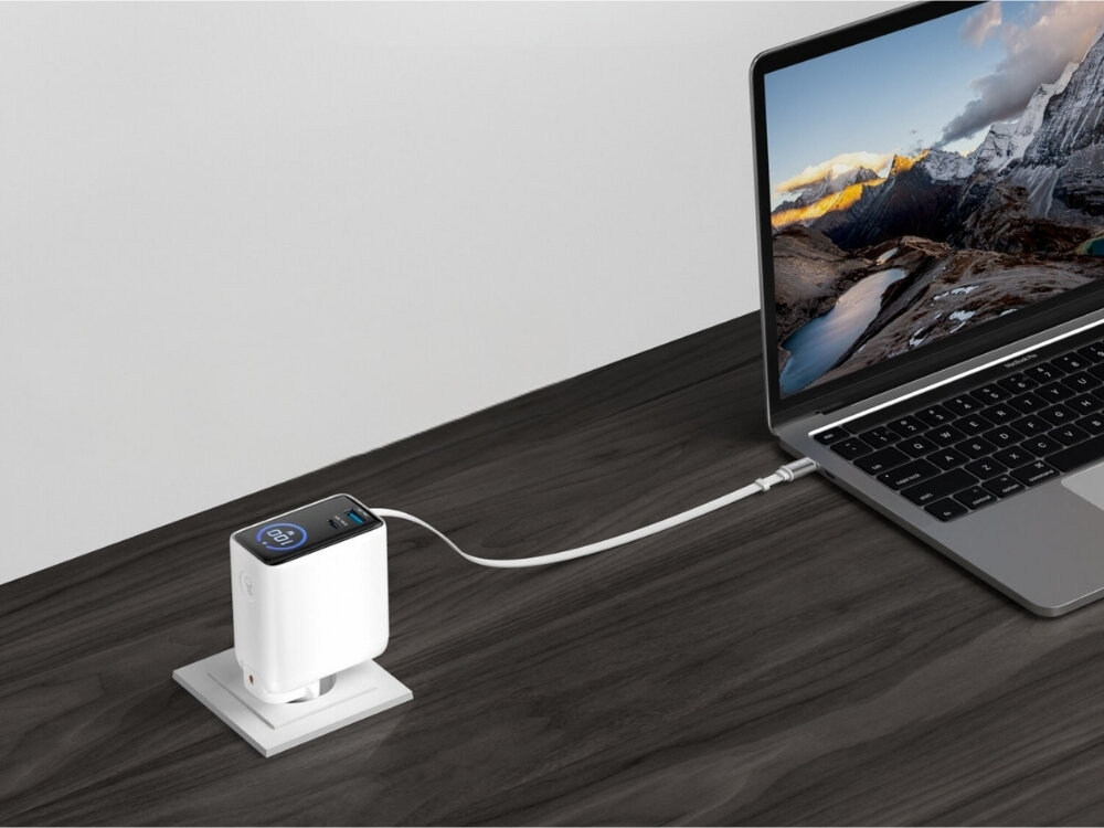 Na drewnianym biurku znajduje się biały adapter sieciowy podłączony do laptopa za pomocą kabla USB-C. Na ekranie laptopa widoczny jest krajobraz górski.