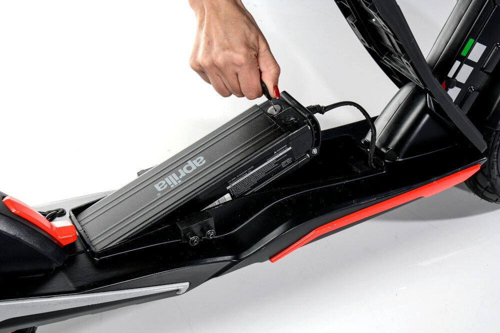 Osoba wkłada baterię do czarno-czerwonej hulajnogi elektrycznej. Na baterii widoczne jest logo \'aprilia\'.