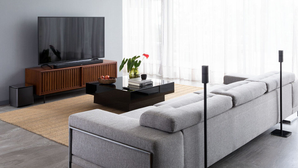 Nowoczesny salon z dużą szarą sofą, telewizorem na drewnianej szafce i stolikiem kawowym z rośliną. W pomieszczeniu znajdują się również głośniki i jasne zasłony na oknach.