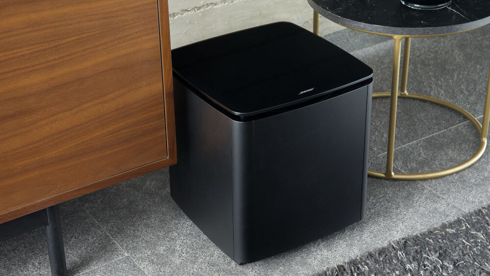 Czarny subwoofer z logo \'Bose\' na górnej powierzchni, ustawiony obok drewnianej szafki i okrągłego stolika z metalowymi nogami.