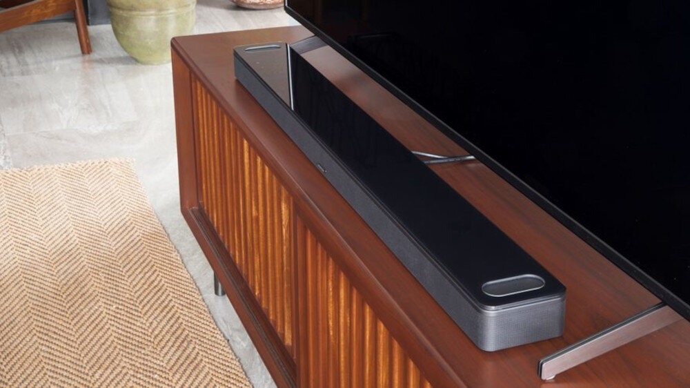 Na drewnianej szafce stoi biały soundbar o nowoczesnym designie. W tle widoczny jest fragment telewizora.