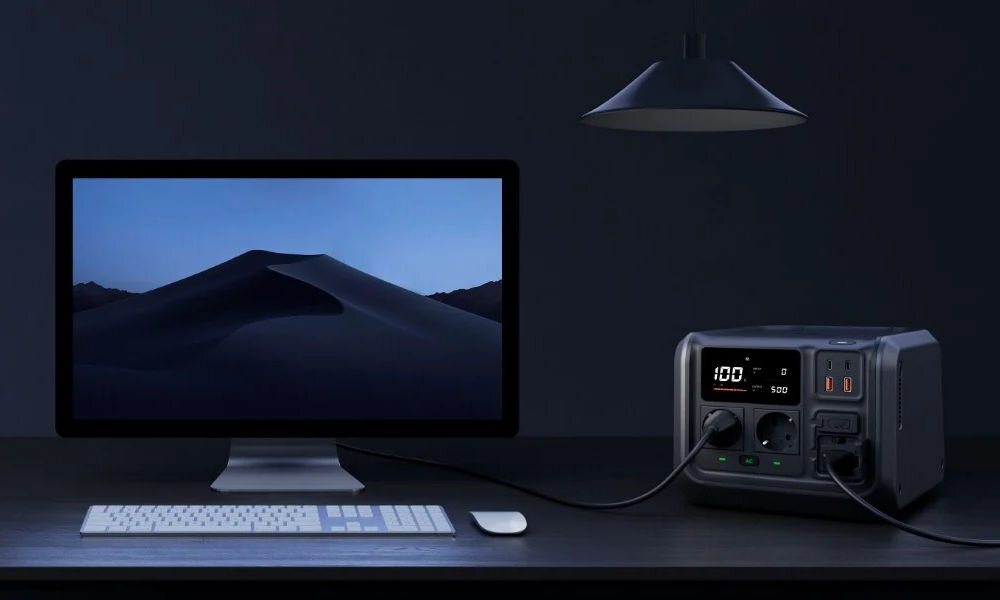 Na biurku znajduje się monitor z wyświetlonym obrazem pustynnego krajobrazu oraz stacja zasilania z wyświetlaczem pokazującym \'100%\'. Obok monitora leży klawiatura i myszka, a nad biurkiem wisi lampa.