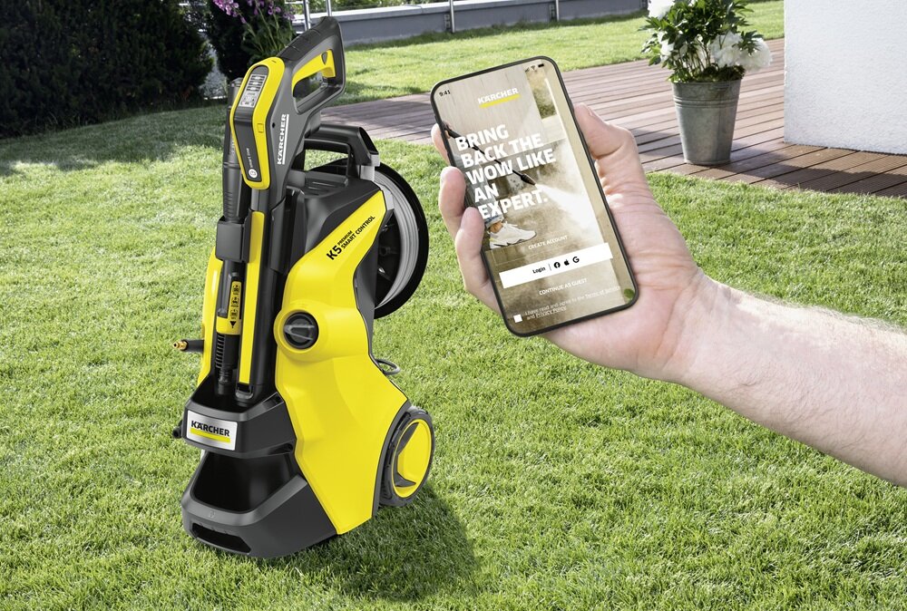 Na trawniku stoi żółto-czarna myjka ciśnieniowa z widocznym logo KARCHER. Obok osoba trzyma smartfon z wyświetlonym tekstem: \'BRING BACK THE WOW LIKE AN EXPERT.\'