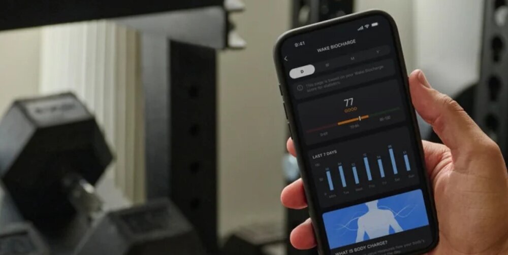 Osoba trzyma smartfon z aplikacją fitness, na ekranie widoczny jest wykres i liczba 77. W tle znajduje się sprzęt do ćwiczeń siłowych.