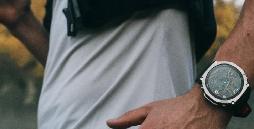 Osoba nosi smartwatch z dużym, okrągłym wyświetlaczem i czarno-srebrną obudową. W tle widoczne są rozmyte drzewa.