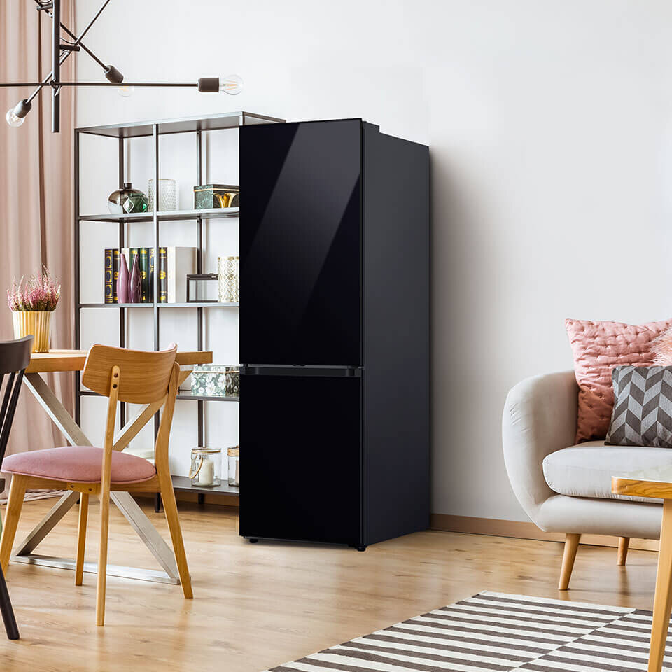 Lodówki Samsung Bespoke występują w dwóch wysokościach: 185 i 203 cm Czarna lodówka o nowoczesnym designie stoi w jasnym, stylowo urządzonym wnętrzu z drewnianą podłogą i meblami. Obok lodówki znajduje się regał z książkami i dekoracjami oraz stół z krzesłami.
