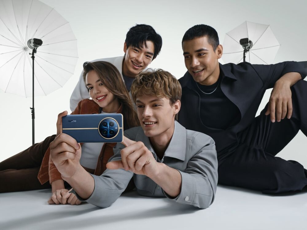 Cztery osoby siedzą razem, jedna z nich trzyma smartfon z widocznym logo \'realme\'. W tle znajdują się dwa duże parasole fotograficzne.