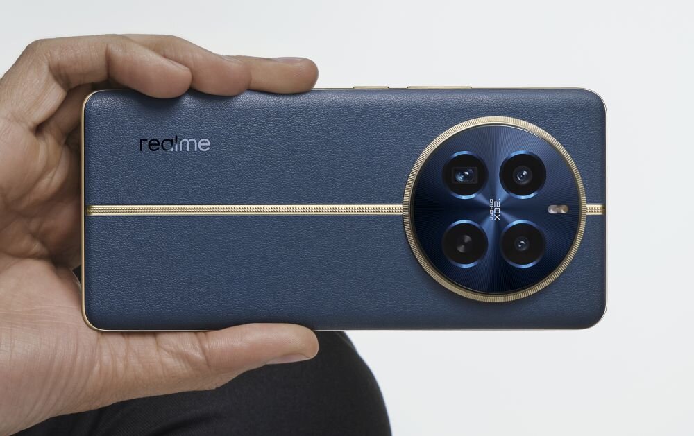 Osoba trzyma smartfon z ciemnoniebieskim tyłem i złotymi akcentami. Na obudowie widoczne jest logo \'realme\' oraz duży okrągły moduł z czterema obiektywami aparatu.