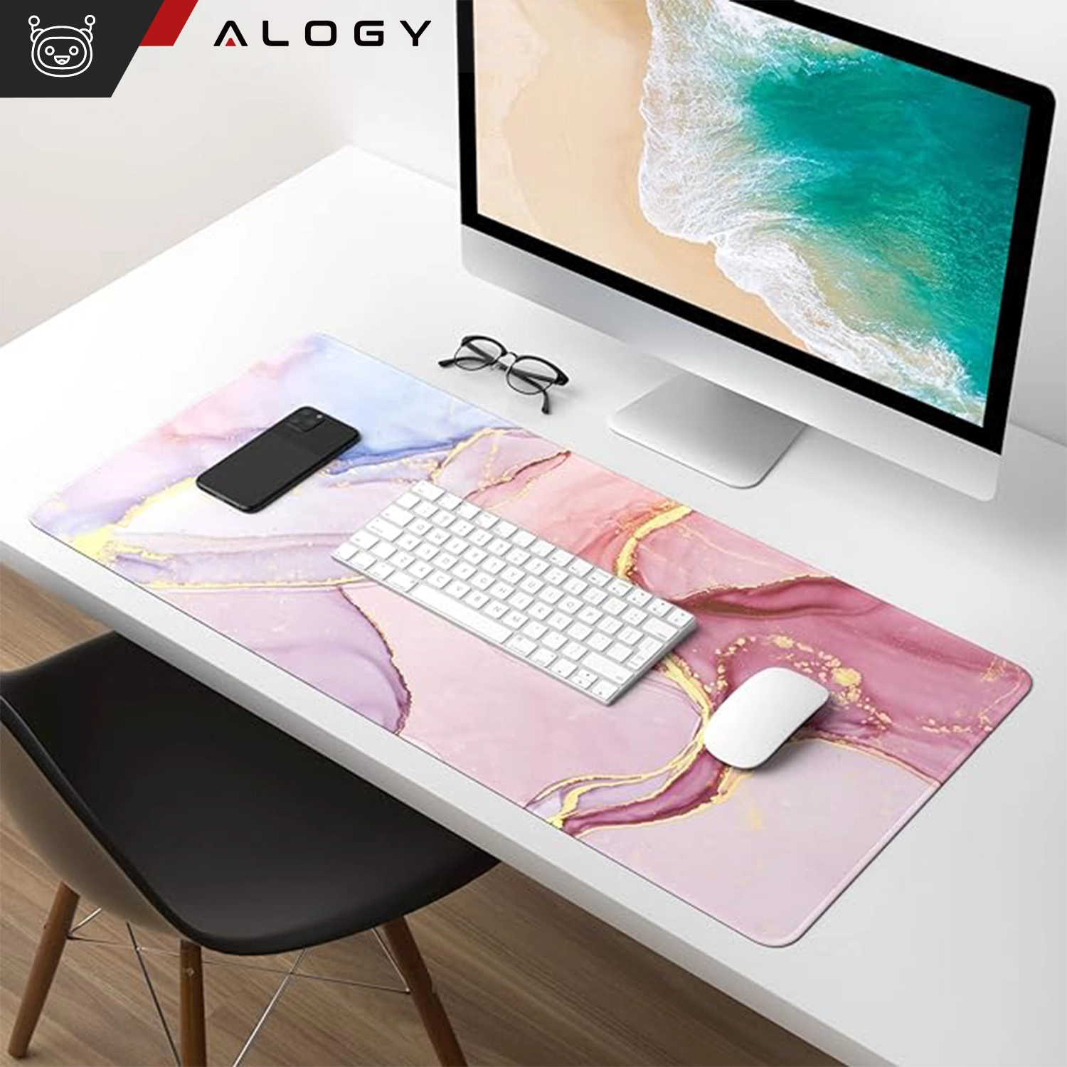 Na biurku znajduje się różowa podkładka z marmurowym wzorem, na której leżą klawiatura, mysz i smartfon. W tle widać monitor z wyświetlonym obrazem plaży oraz logo \'Alogy\'.