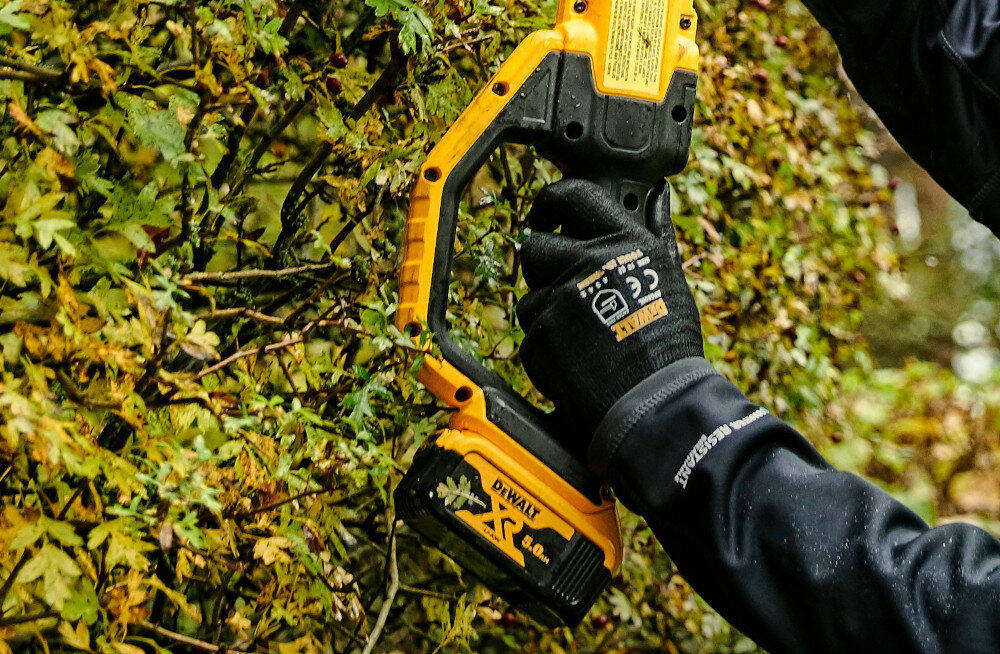 Osoba trzyma akumulatorowe nożyce do żywopłotu w kolorze żółto-czarnym, przycinając zielony żywopłot. Na urządzeniu widoczne jest logo \'DEWALT\' oraz napis \'XR 5.0Ah\'.