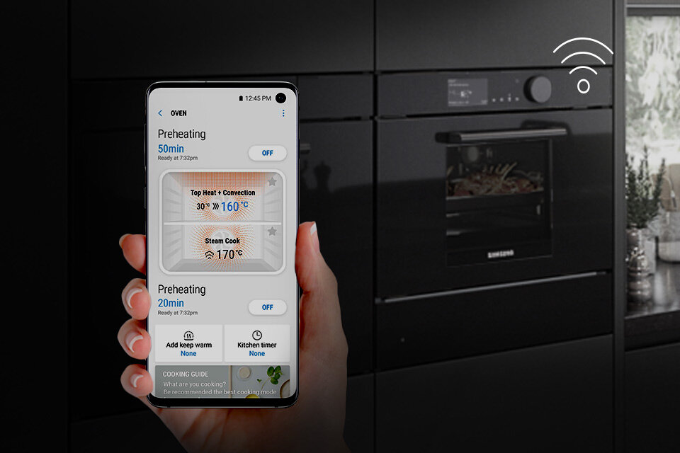 Osoba trzyma smartfon z aplikacją do sterowania piekarnikiem, na ekranie widoczne są ustawienia temperatury i czasu pieczenia. W tle znajduje się czarny piekarnik z logo Samsung.
