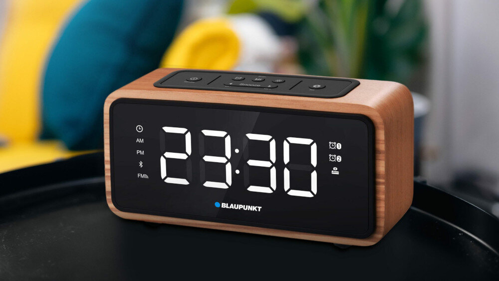 Radiobudzik z drewnianą obudową i czarnym frontem wyświetla godzinę 23:30 na dużym, białym wyświetlaczu LED. Na górze urządzenia znajdują się przyciski sterujące, a poniżej wyświetlacza widoczne jest logo BLAUPUNKT.