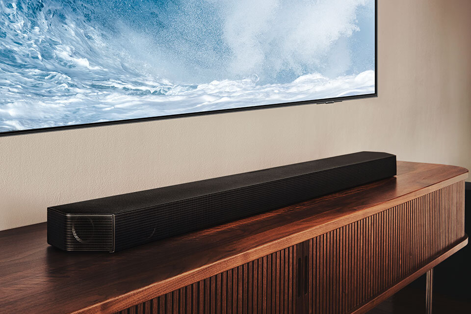 Na drewnianej szafce stoi czarny soundbar, a nad nim wisi telewizor wyświetlający obraz fal morskich.