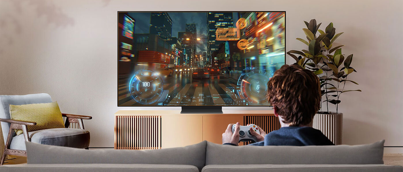 Samsung OLED S95C to telewizor z szeregiem udogodnień dla graczy Osoba siedzi na kanapie przed dużym telewizorem, na którym wyświetlana jest dynamiczna scena z gry wyścigowej. W tle widać roślinę doniczkową i minimalistyczny stolik RTV.