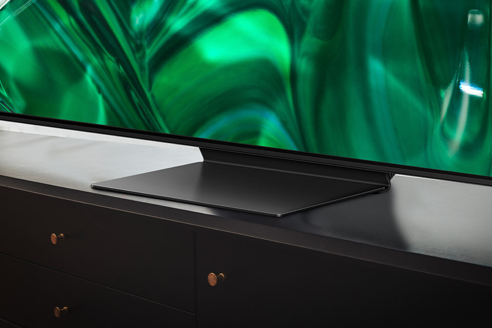Telewizor z cienką ramką stoi na ciemnej szafce, a na ekranie widoczny jest zielony, abstrakcyjny wzór.