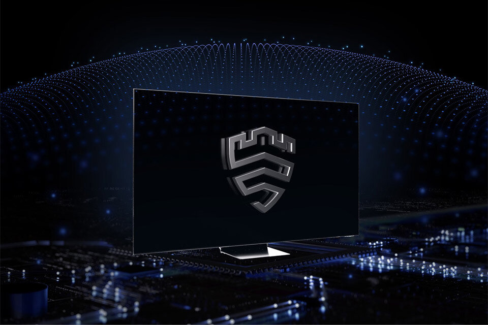 Samsung Knox Na obrazie widoczny jest telewizor z dużym ekranem, na którym wyświetlone jest logo w kształcie tarczy. W tle widać abstrakcyjne wzory świetlne na ciemnym tle.