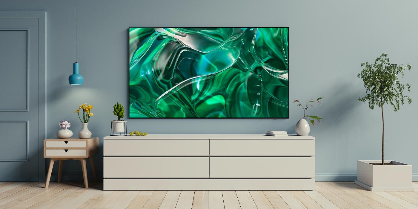 phpwg3smp ls 01 1400 Na ścianie wisi duży telewizor z wyświetlonym abstrakcyjnym, zielonym wzorem. Przed telewizorem znajduje się niska szafka z kilkoma dekoracjami, a obok stoi roślina w doniczce.