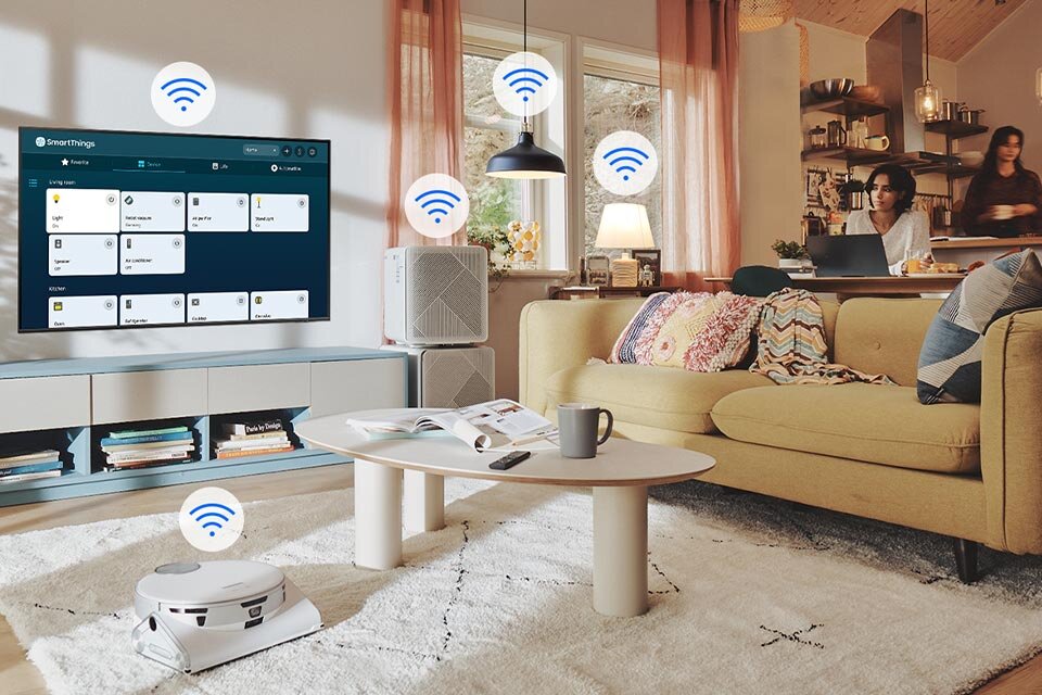 W salonie znajduje się telewizor z wyświetlonym interfejsem \'SmartThings\', obok którego widoczne są ikony sygnału Wi-Fi. Na stoliku leży otwarta książka i kubek, a w tle widać dwie osoby przy stole z laptopem.
