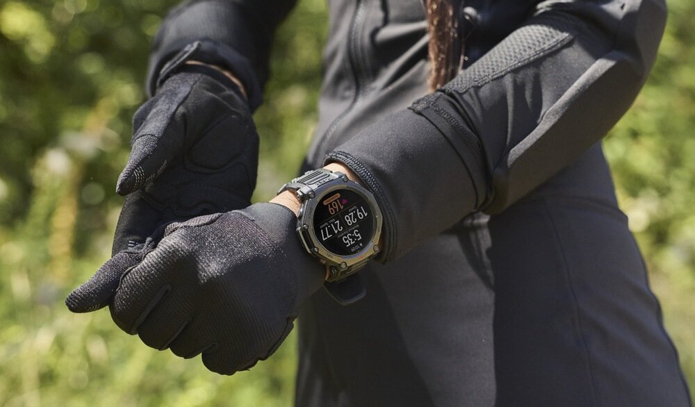 Osoba w czarnych rękawicach i odzieży sportowej nosi smartwatch z okrągłym wyświetlaczem, na którym widoczna jest godzina 5:35 oraz data 19/28 12:11. W tle znajduje się rozmyta zieleń.