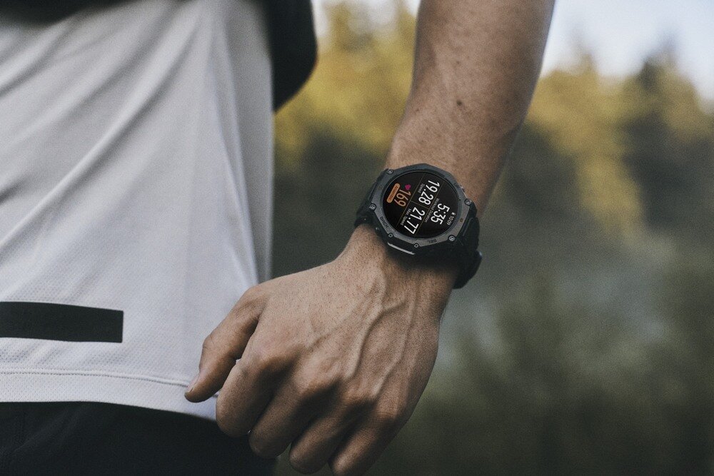 Osoba nosi smartwatch na nadgarstku, wyświetlający dane takie jak czas i liczba kroków. W tle widoczne są rozmyte drzewa.