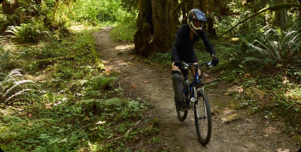 Osoba na rowerze górskim jedzie ścieżką w lesie, otoczoną bujną zielenią i drzewami. Osoba ma na sobie kask i ochraniacze.
