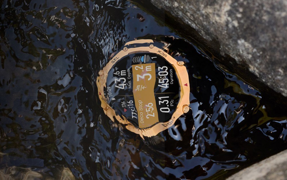 Smartwatch w złotym kolorze zanurzony w wodzie, wyświetlający dane takie jak głębokość 43 m, czas 45:03 i inne parametry. Na ekranie widoczny jest tekst: \'Deco Stop 2:56\'.