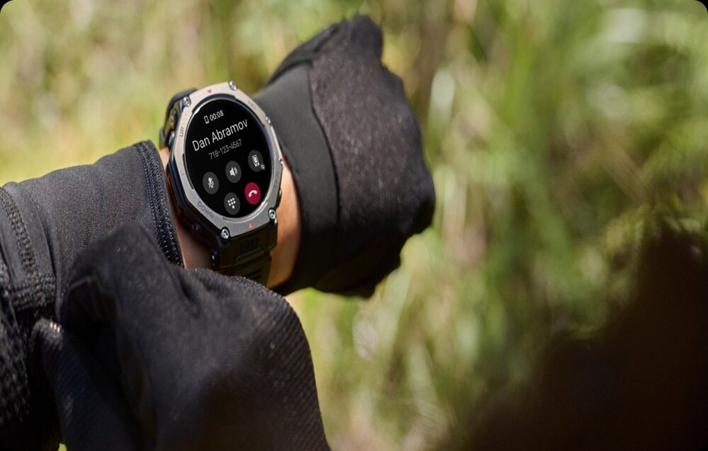 Osoba nosi smartwatch z wyświetlaczem pokazującym połączenie przychodzące od \'Dan Abramov\'. W tle widoczna jest rozmyta zieleń.
