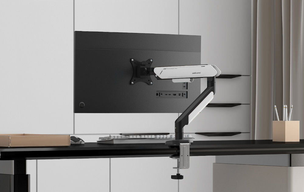 Na biurku zamontowany jest biało-czarny uchwyt do monitora z logo \'anda seat\'. W tle widoczny jest monitor oraz zasłony.
