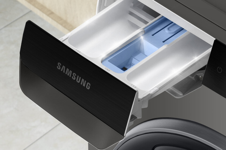 System przepłukiwania wodą Obraz przedstawia otwarty pojemnik na detergenty pralko-suszarki z widocznym logo \'SAMSUNG\'. Wnętrze pojemnika jest białe z niebieskim elementem w środku.