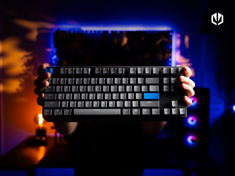 Osoba trzyma klawiaturę mechaniczną z podświetleniem, na której kilka klawiszy jest w kolorze niebieskim. W tle widoczne są kolorowe światła oraz logo ENDORFY.