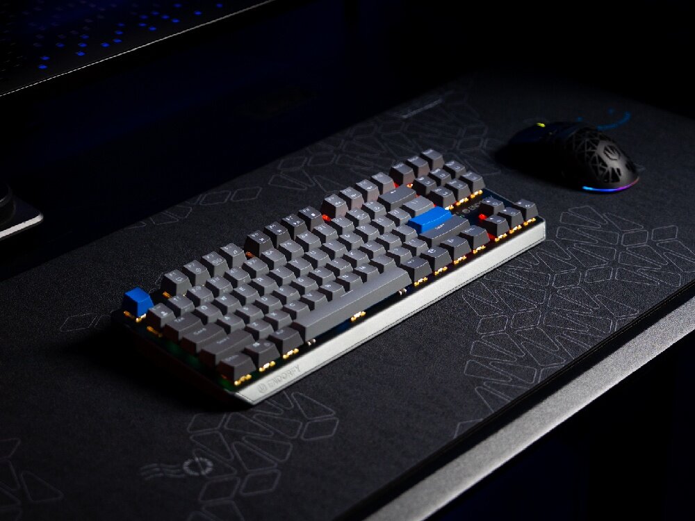 Na ciemnym biurku znajduje się klawiatura mechaniczna z szarymi klawiszami i niebieskimi akcentami. Obok klawiatury leży mysz komputerowa z podświetleniem RGB.