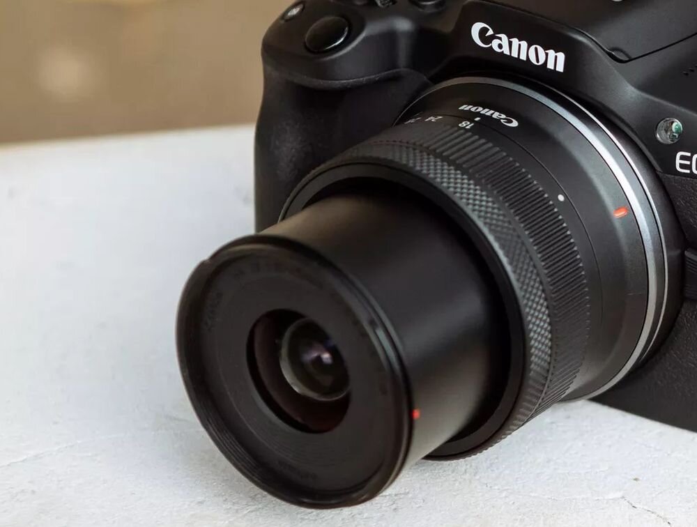 Zbliżenie na aparat bezlusterkowy z obiektywem, na którym widoczne jest logo \'Canon\'. Obiektyw ma oznaczenia \'18-45mm\'.