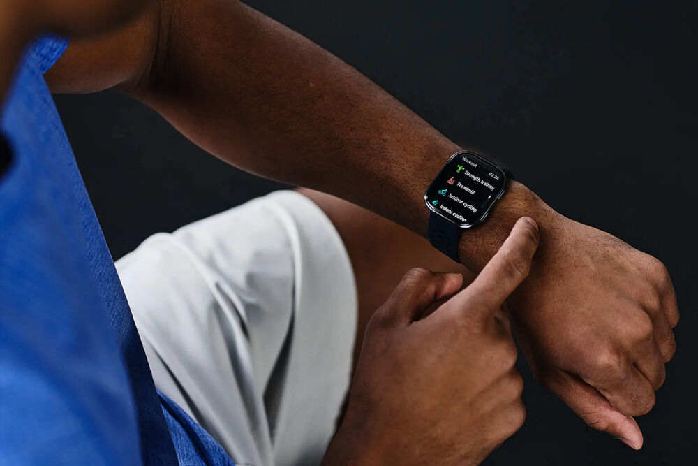 Osoba nosi czarny smartwatch na nadgarstku, na ekranie widoczne są różne ikony i tekst. Tło jest ciemne, a osoba ubrana jest w niebieską koszulę i jasne spodnie.