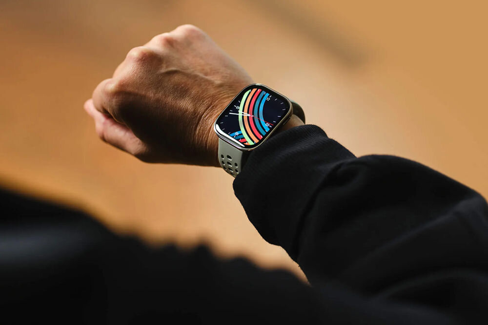 Osoba nosi smartwatch z kolorowym wyświetlaczem na nadgarstku. Pasek zegarka jest jasny z otworami wentylacyjnymi.