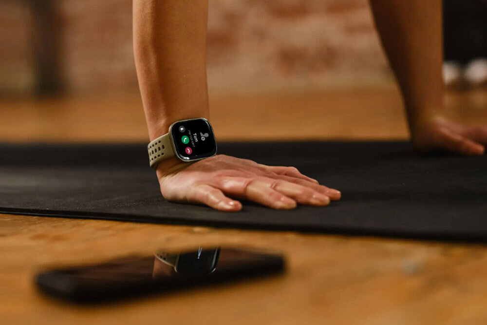 Osoba wykonuje ćwiczenie na macie, nosząc smartwatch na nadgarstku. Na podłodze leży smartfon.