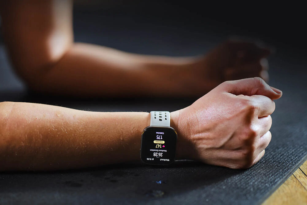 Osoba leży na macie, na nadgarstku ma założony smartwatch z wyświetlaczem pokazującym dane dotyczące tętna i kroków. Pasek zegarka jest jasny z otworami wentylacyjnymi.