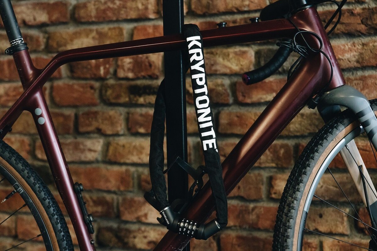 Na tle ceglanej ściany widać rower z przypiętym zapięciem rowerowym z napisem \'KRYPTONITE\'. Zapięcie ma mechanizm szyfrowy z czterema pokrętłami.