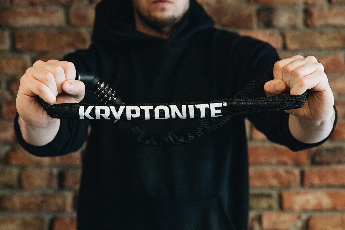 Osoba trzyma zapięcie rowerowe z widocznym napisem \'KRYPTONITE\' na tle ceglanej ściany.