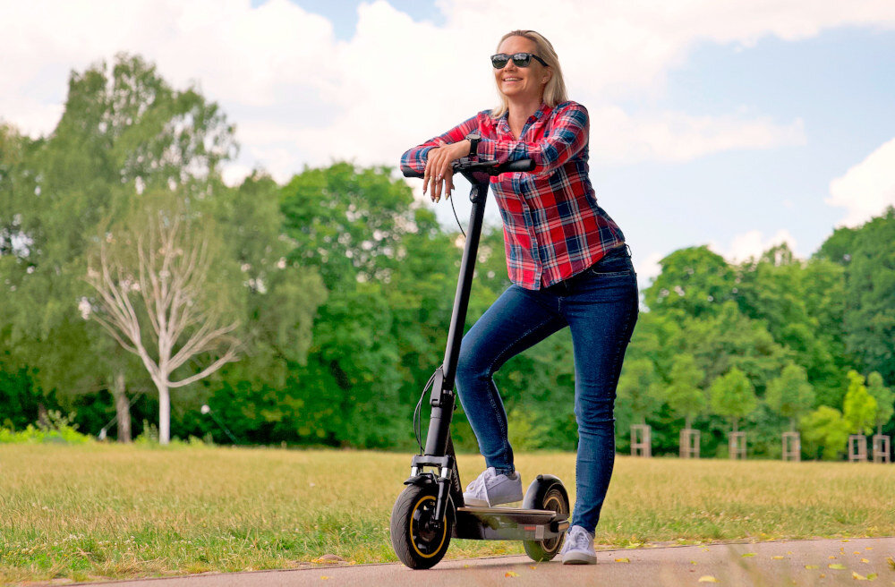 Osoba w okularach przeciwsłonecznych stoi na ścieżce, opierając się o czarną hulajnogę elektryczną. W tle widoczny jest zielony park z drzewami i trawnikiem.