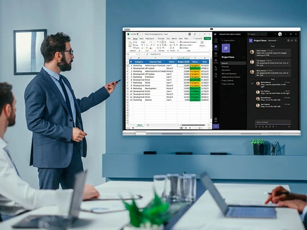 Mężczyzna w garniturze wskazuje na duży monitor, na którym wyświetlane są arkusz kalkulacyjny i aplikacja do komunikacji. W tle widać nowoczesne wnętrze biurowe z osobą pracującą na laptopie.