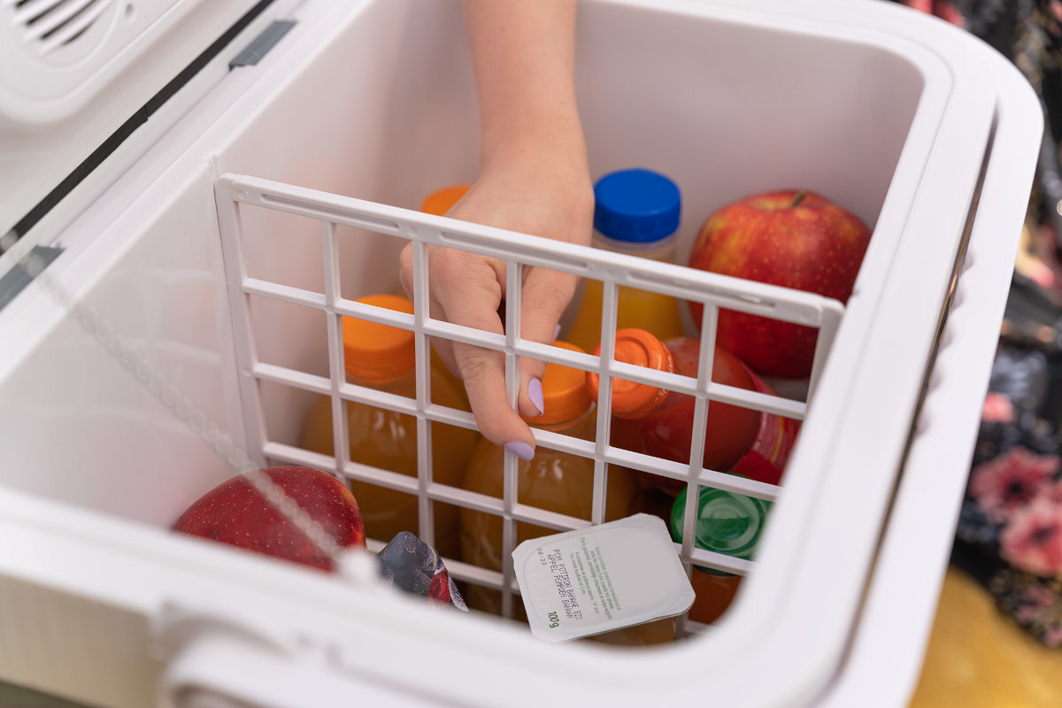 Wnętrze lodówki turystycznej z widocznymi butelkami napojów, jabłkiem i ręką sięgającą po jeden z produktów. W środku znajduje się również kratka oddzielająca produkty.