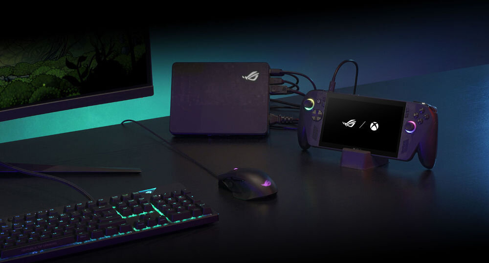 Na biurku znajduje się konsola z ekranem, na którym widoczne są loga ROG i Xbox, obok klawiatura, myszka oraz urządzenie z logo ROG. Wszystkie elementy są podłączone kablami.